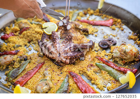 Seafood paella with whole octopus, peppers, lemon slices being served from metal pan at food festival. Concept of seafood, exotic dish, gourmet cooking on display Seafood paella with whole octopus, peppers, lemon slices being served from metal pan at food festival. Concept of seafood, exotic dish, gourmet cooking on display 127652035