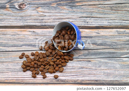 Close-up and top view of hot black coffee in blue coffee cup and roasted Thai coffee beans on wooden background. Close-up and top view of hot black coffee in blue coffee cup and roasted Thai coffee beans on wooden background. 127652097