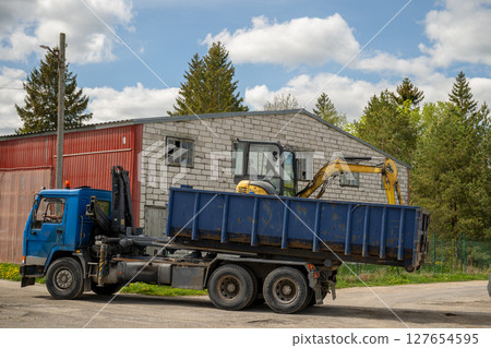 Blue industrial truck carrying a yellow mini excavator in its container bed 127654595