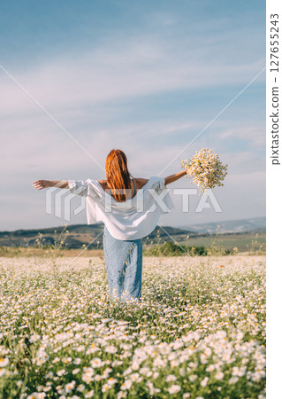 Daisies Woman Meadow: Redhead spreads arms holding flowers in daisy field during daytime. Daisies Woman Meadow: Redhead spreads arms holding flowers in daisy field during daytime. 127655243