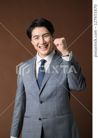 The upper body of a businessman in a suit pumping his fist against a brown background. Image of a professional or real estate executive 127655870