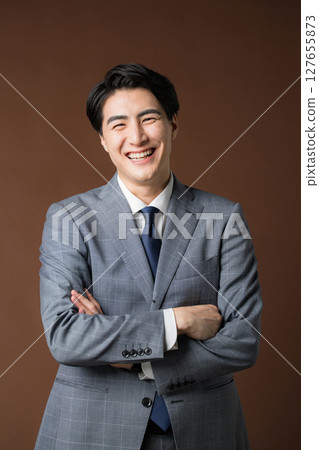 一位憂心忡忡、深思的商人總統身穿西裝,雙臂交叉,背景為棕色,面帶微笑 一位憂心忡忡、深思的商人總統身穿西裝,雙臂交叉,背景為棕色,面帶微笑 127655873