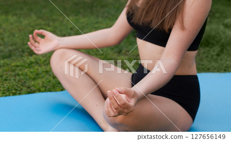 Young woman in black sportswear sitting on a yoga mat in lotus position, meditating outdoors on green grass 127656149