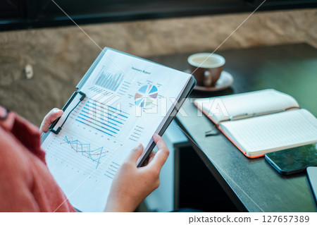 Close-up of a person reviewing financial charts and graphs, with a notebook and coffee on a desk. Close-up of a person reviewing financial charts and graphs, with a notebook and coffee on a desk. 127657389