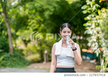 A woman takes a break during her workout, drinking water in a vibrant green park. Fitness and hydration concept. 127657418