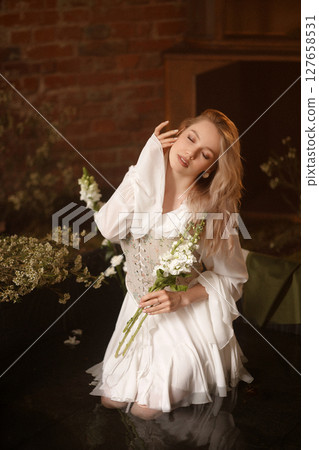 Young woman in white dress sitting in artificial pond with flowers and fog against brick wall Young woman in white dress sitting in artificial pond with flowers and fog against brick wall 127658531