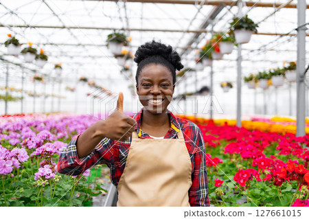 A smiling African woman gives a thumbs-up in a greenhouse filled with vibrant flowers, showing positivity and approval. A smiling African woman gives a thumbs-up in a greenhouse filled with vibrant flowers, showing positivity and approval. 127661015
