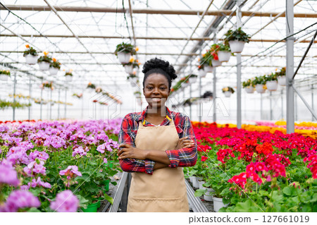 A smiling African woman stands in a greenhouse filled with colorful flowers, arms crossed. A smiling African woman stands in a greenhouse filled with colorful flowers, arms crossed. 127661019