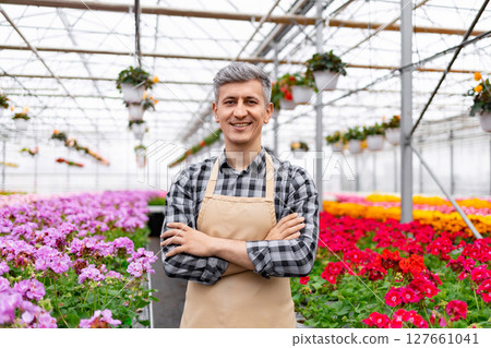 A smiling man in an apron stands in a greenhouse, surrounded by colorful flowers. 127661041