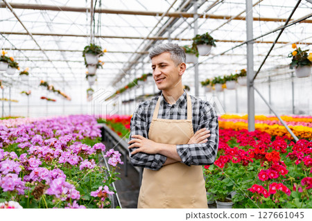 A smiling man with crossed arms stands in a sunlit greenhouse, surrounded by vibrant flowers and hanging plants. A smiling man with crossed arms stands in a sunlit greenhouse, surrounded by vibrant flowers and hanging plants. 127661045