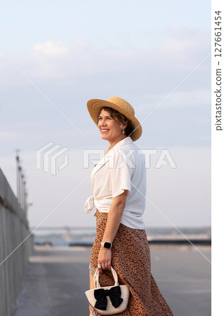 Smiling woman wearing straw hat walks outdoors at sunset. 127661454