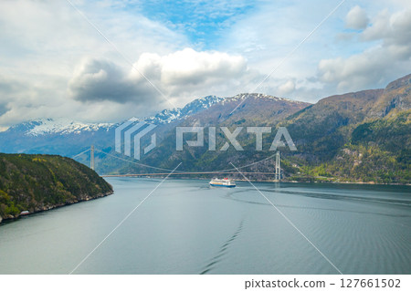 Distant tourist ship sails under Hardanger Bridge gracefully spanning waters of fjord. Mountains with snow on tops create Norwegian landscape 127661502
