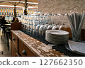 Close-up of clean glasses, utensils, stacked plates on weathered wooden countertop at outdoor cafe, Lisbon, Portugal. Relaxed dining atmosphere, people seated, umbrellas, historic city square, travel 127662350
