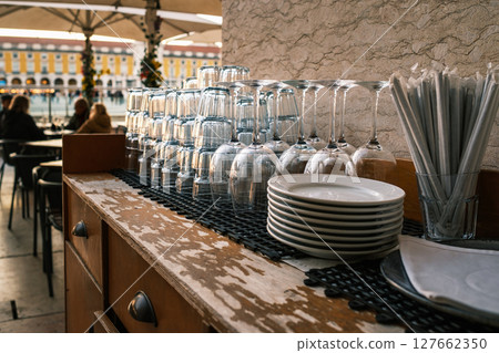 Close-up of clean glasses, utensils, stacked plates on weathered wooden countertop at outdoor cafe, Lisbon, Portugal. Relaxed dining atmosphere, people seated, umbrellas, historic city square, travel Close-up of clean glasses, utensils, stacked plates on weathered wooden countertop at outdoor cafe, Lisbon, Portugal. Relaxed dining atmosphere, people seated, umbrellas, historic city square, travel 127662350