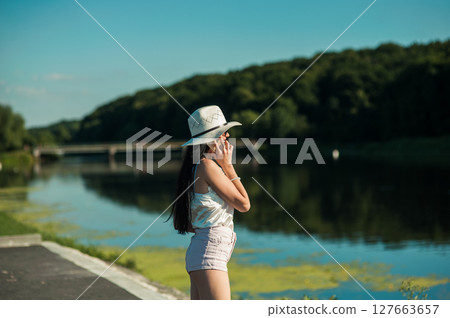 A slender girl by the water, dressed in a T-shirt and shorts, holds a phone and talks 127663657