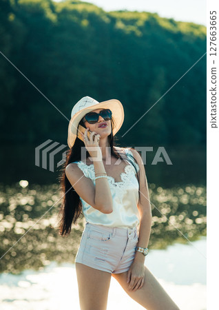 A girl in a white hat is standing by the pond and smiling sincerely while holding the phone A girl in a white hat is standing by the pond and smiling sincerely while holding the phone 127663665