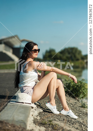 A young woman in a light outfit is sitting comfortably on the ground, catching the warmth of the sun A young woman in a light outfit is sitting comfortably on the ground, catching the warmth of the sun 127663672