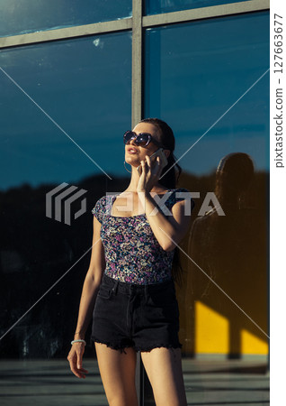 A girl wearing a light T-shirt and shorts is standing next to a glass building, holding a cell phone to her ear A girl wearing a light T-shirt and shorts is standing next to a glass building, holding a cell phone to her ear 127663677