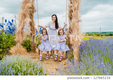 A young mother gently swings her twin daughters on a large, picturesque swing A young mother gently swings her twin daughters on a large, picturesque swing 127665210