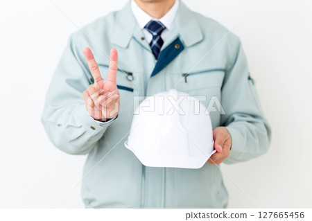 A middle-aged male businessman wearing green work clothes, holding a helmet and making a peace sign 127665456