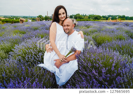 The man in a white T-shirt embraces his wife, who is sitting on his lap in the lavender field The man in a white T-shirt embraces his wife, who is sitting on his lap in the lavender field 127665695