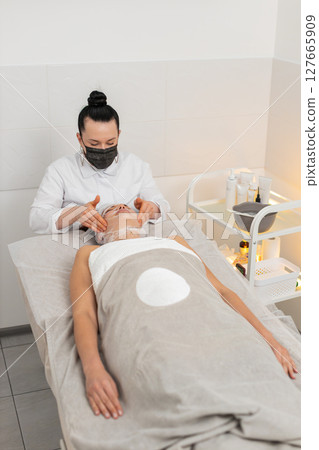 Woman completing facial wrap mask procedure in cosmetology clinic, beautician apply gauze skin care 127665909