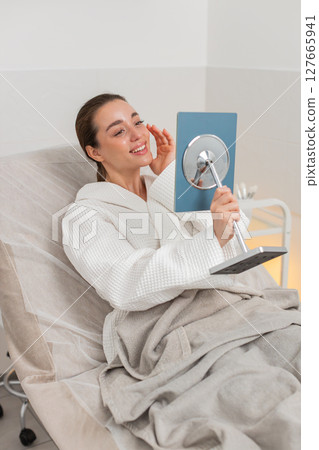 Caucasian young woman smiles in mirror after facial therapy in beauty clinic showing visible result Caucasian young woman smiles in mirror after facial therapy in beauty clinic showing visible result 127665941