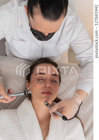 Caucasian young woman gets lip zone facial therapy with modern device by cosmetologist in clinic Caucasian young woman gets lip zone facial therapy with modern device by cosmetologist in clinic 127665942