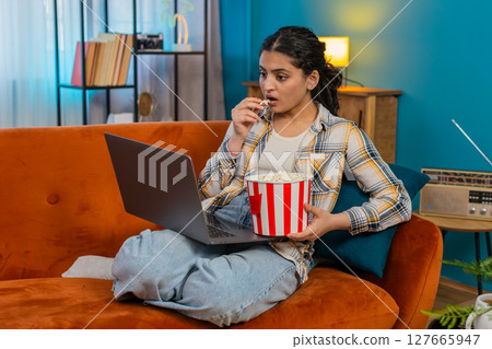 Indian woman sitting on sofa watching movie on laptop laughing loudly lively reaction eating popcorn Indian woman sitting on sofa watching movie on laptop laughing loudly lively reaction eating popcorn 127665947