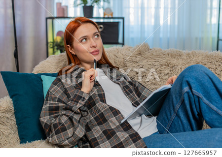 Woman sitting on sofa writing thoughtful notes in journal dreaming and planning with calm expression Woman sitting on sofa writing thoughtful notes in journal dreaming and planning with calm expression 127665957