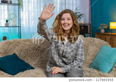 Woman sitting on sofa smiling and waving hand hello with joyful face greeting in friendly manner 127665958