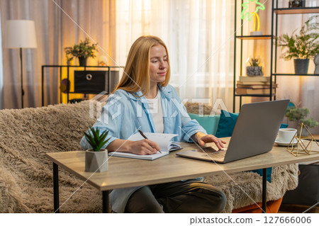 Young woman studying at home video laptop lesson focus, notes in notebook, attentive learning moment 127666006