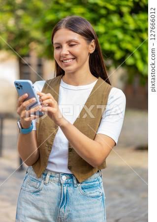 Smiling young woman using smartphone typing texting scrolling social media app on city town street Smiling young woman using smartphone typing texting scrolling social media app on city town street 127666012