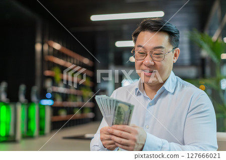 Smiling mature Chinese businessman in formal shirt counting dollar banknotes in modern office lobby Smiling mature Chinese businessman in formal shirt counting dollar banknotes in modern office lobby 127666021