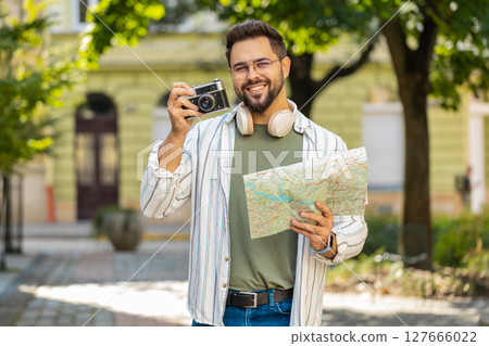 Happy young man traveler tourist holding map and taking photos using retro camera on city street Happy young man traveler tourist holding map and taking photos using retro camera on city street 127666022