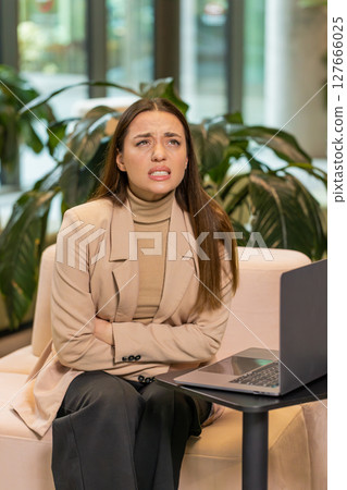 Disappointed Caucasian businesswoman using laptop and feeling sudden stomach ache in office lobby Disappointed Caucasian businesswoman using laptop and feeling sudden stomach ache in office lobby 127666025