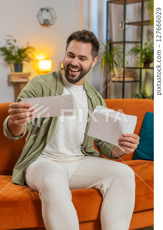 Excited young man opening envelope letter and reading good news sitting on sofa at home apartment 127666029