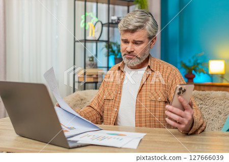 Mature man at table with laptop and smartphone reviewing financial reports, showing exhaustion 127666039