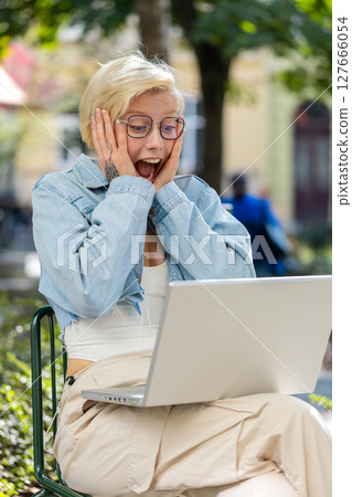 Overjoyed happy young woman working on laptop screaming in delight raises hands in city street 127666054