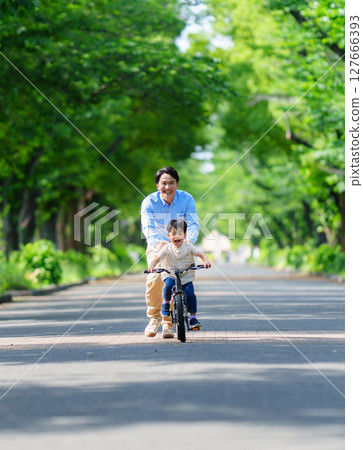 A 5-year-old boy practices riding a bicycle with his father following him A 5-year-old boy practices riding a bicycle with his father following him 127666393