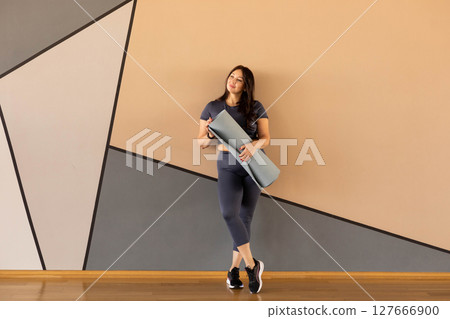 Curvy woman in activewear leaning on wall with yoga mat, smiling before workout 127666900