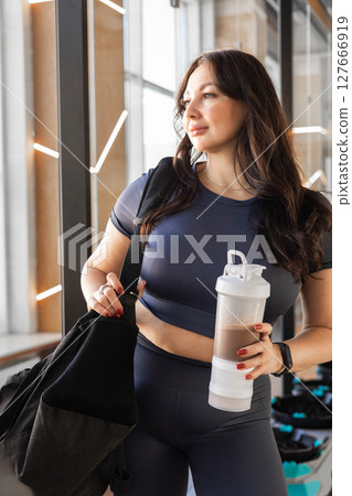 Curvy brunette woman in gym outfit holding sports bag and protein drink at modern fitness studio Curvy brunette woman in gym outfit holding sports bag and protein drink at modern fitness studio 127666919