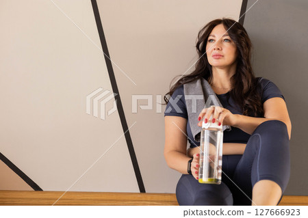 Fit woman in sportswear resting after workout with water bottle, sitting and smiling in gym interior 127666923