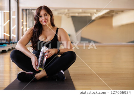 Plus-size woman sitting on mat at gym holding water bottle after workout, body positivity 127666924