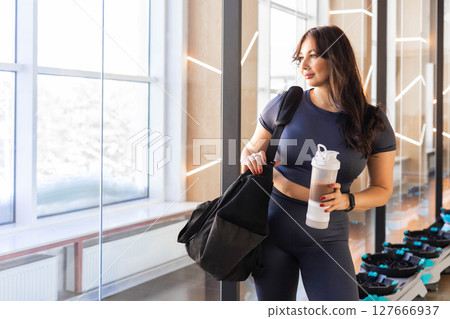 Curvy woman in gym wear holding a protein shaker by window, fitness studio scene, full body Curvy woman in gym wear holding a protein shaker by window, fitness studio scene, full body 127666937