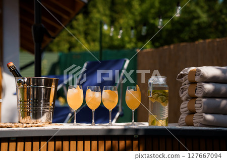 Luxurious Summer Patio Scene with Champagne, Cocktails, Towels, and Outdoor Relaxation Setup. Golden Hour, Wooden Deck, Modern Furniture. 127667094