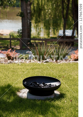 Tranquil Backyard Scene with Round Fire Pit and Lush Greenery Featuring a Lake View in Soft Evening Light, Outdoor Living Relaxation 127667099