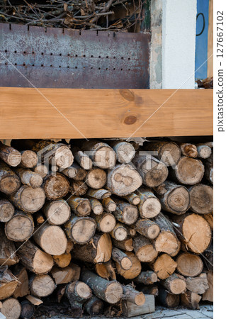 Rustic Woodpile with Log Stack, Weathered Metal Panel, and Light Wood Beam. Outdoor Firewood Storage Scene in Natural Brown Tones and Textures. Rustic Woodpile with Log Stack, Weathered Metal Panel, and Light Wood Beam. Outdoor Firewood Storage Scene in Natural Brown Tones and Textures. 127667102