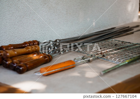Rustic BBQ Tools Laid Out on Stone Countertop. Grills, Skewers, Tongs, Wood Handles, Metal Texture, Brown and Gray Culinary Still Life. Rustic BBQ Tools Laid Out on Stone Countertop. Grills, Skewers, Tongs, Wood Handles, Metal Texture, Brown and Gray Culinary Still Life. 127667104