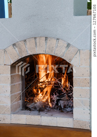 Rustic Outdoor Fireplace Burning Wood with Arched Opening: Textured Stone Hearth, Warm Flames, Cozy Backyard Scene, Sunset Glow, Relaxing Ambiance. Rustic Outdoor Fireplace Burning Wood with Arched Opening: Textured Stone Hearth, Warm Flames, Cozy Backyard Scene, Sunset Glow, Relaxing Ambiance. 127667108